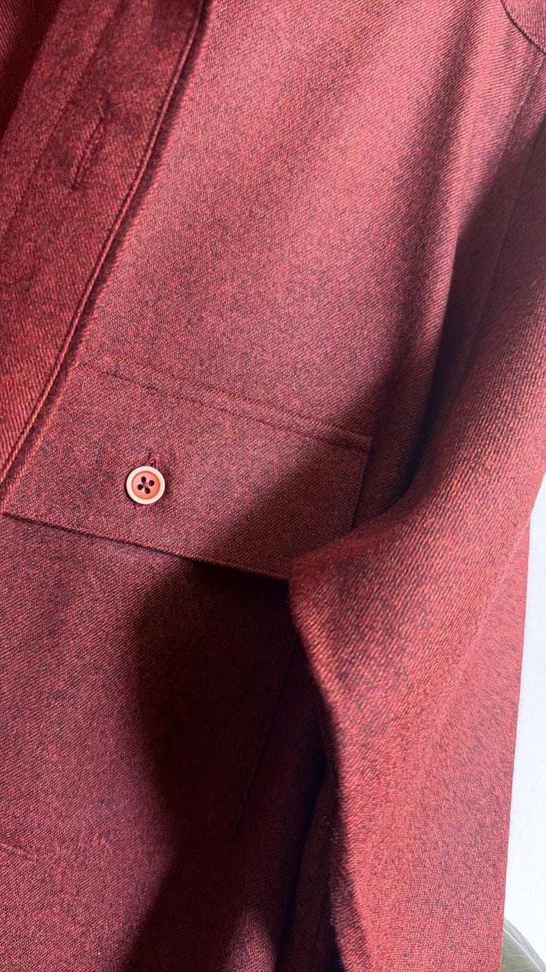 May include: A long-sleeved shirt in a deep burgundy red color with a textured, heathered fabric finish. The shirt features a front chest pocket with a single, centered, light-colored button that has four small holes in the middle of the button.