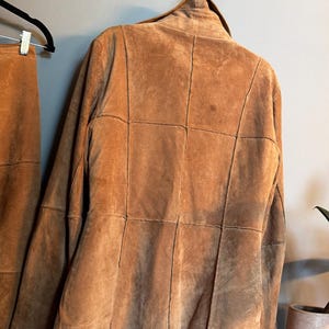 May include: A tan suede jacket featuring a panelled construction with visible vertical and horizontal seams across the back. The jacket has a structured collar and long sleeves, presented hanging on a black hanger against a plain wall.