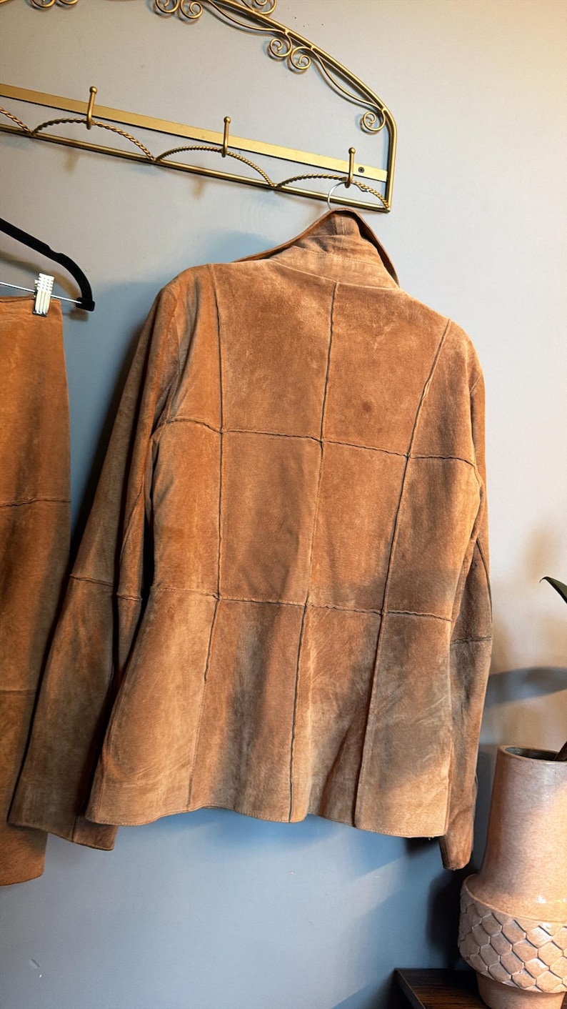 May include: A tan suede jacket featuring a panelled construction with visible vertical and horizontal seams across the back. The jacket has a structured collar and long sleeves, presented hanging on a black hanger against a plain wall.