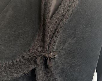 May include: A black suede jacket featuring a dark brown cable-knit shawl collar and hem trim. The jacket has a single button closure at the front and a label inside the collar that reads Coldwater Creek. The garment is displayed on a black hanger.