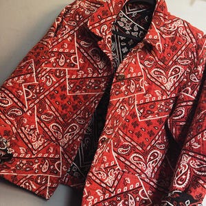 May include: A red and white bandana-print jacket featuring a classic paisley pattern throughout. The garment has a collared neckline, long sleeves with folded cuffs, and a button-down front closure. The interior lining is black with a white paisley print.