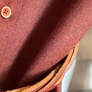 May include: A deep maroon fabric garment featuring a single round button with a light-colored rim. A braided brown leather strap or belt is wrapped around the fabric, showing a textured, layered appearance with visible stitching and edges.