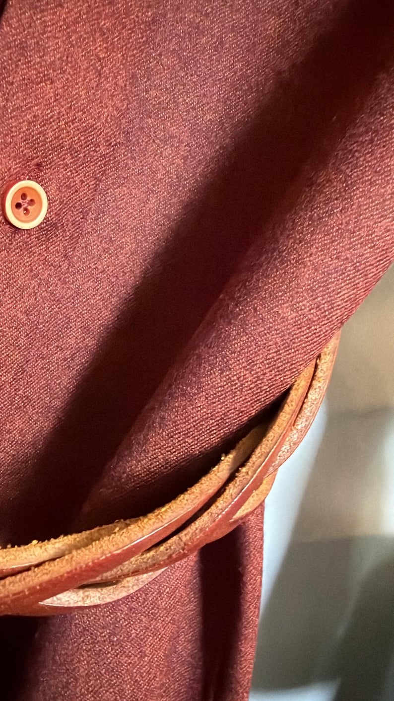 May include: A deep maroon fabric garment featuring a single round button with a light-colored rim. A braided brown leather strap or belt is wrapped around the fabric, showing a textured, layered appearance with visible stitching and edges.