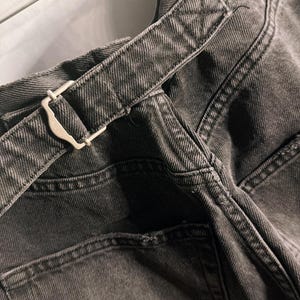 May include: Close-up of dark gray denim pants with a matching belt. The belt features a silver-colored buckle. The pants have a classic five-pocket design and visible stitching details. The fabric appears to be a medium weight denim.