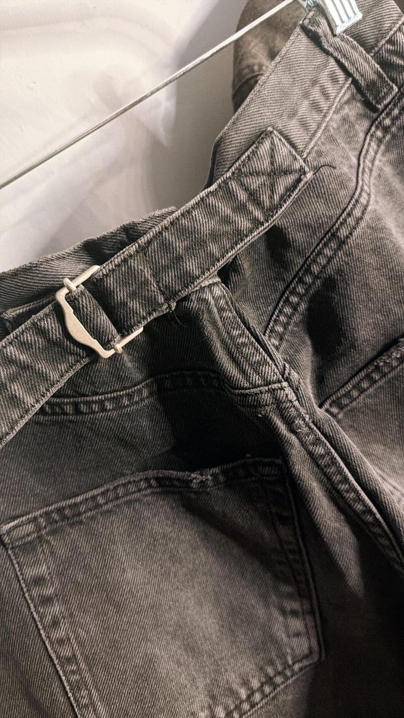 May include: Close-up of dark gray denim pants with a matching belt. The belt features a silver-colored buckle. The pants have a classic five-pocket design and visible stitching details. The fabric appears to be a medium weight denim.