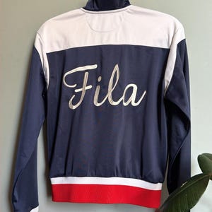 Op de afbeelding: Marineblauwe en witte trainingsjas met rode accenten. De jas heeft het woord "Fila" in witte letters op de rug geborduurd. Het heeft een hoge kraag en hangt aan een goudkleurige wandhaak.
