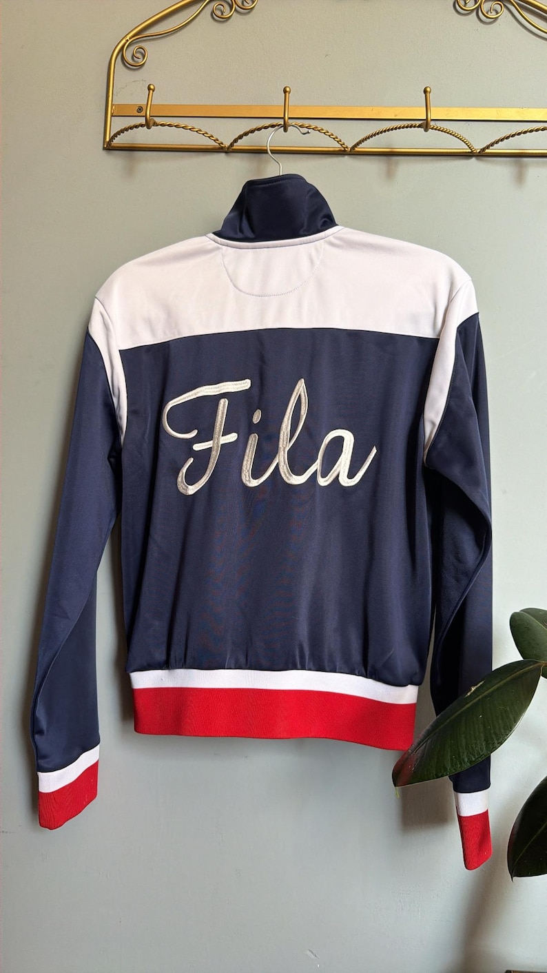 Op de afbeelding: Marineblauwe en witte trainingsjas met rode accenten. De jas heeft het woord "Fila" in witte letters op de rug geborduurd. Het heeft een hoge kraag en hangt aan een goudkleurige wandhaak.