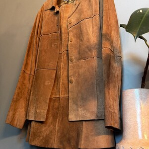 May include: A tan suede jacket featuring a collared neckline, long sleeves, and a front button closure. The garment is constructed from multiple rectangular panels of suede leather, creating a visible grid-like pattern across the entire surface.