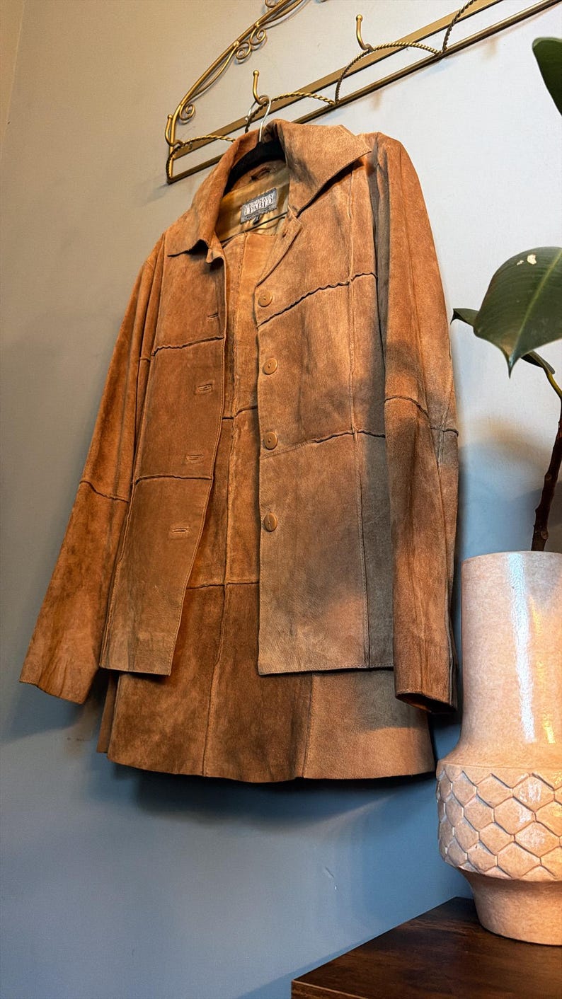 May include: A tan suede jacket featuring a collared neckline, long sleeves, and a front button closure. The garment is constructed from multiple rectangular panels of suede leather, creating a visible grid-like pattern across the entire surface.