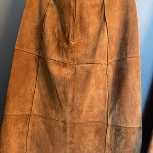 May include: A tan suede skirt featuring a panelled construction with visible vertical and horizontal seams. The skirt has a centred vertical zip closure at the top and is suspended from a black hanger with two metal clips at the waistband.