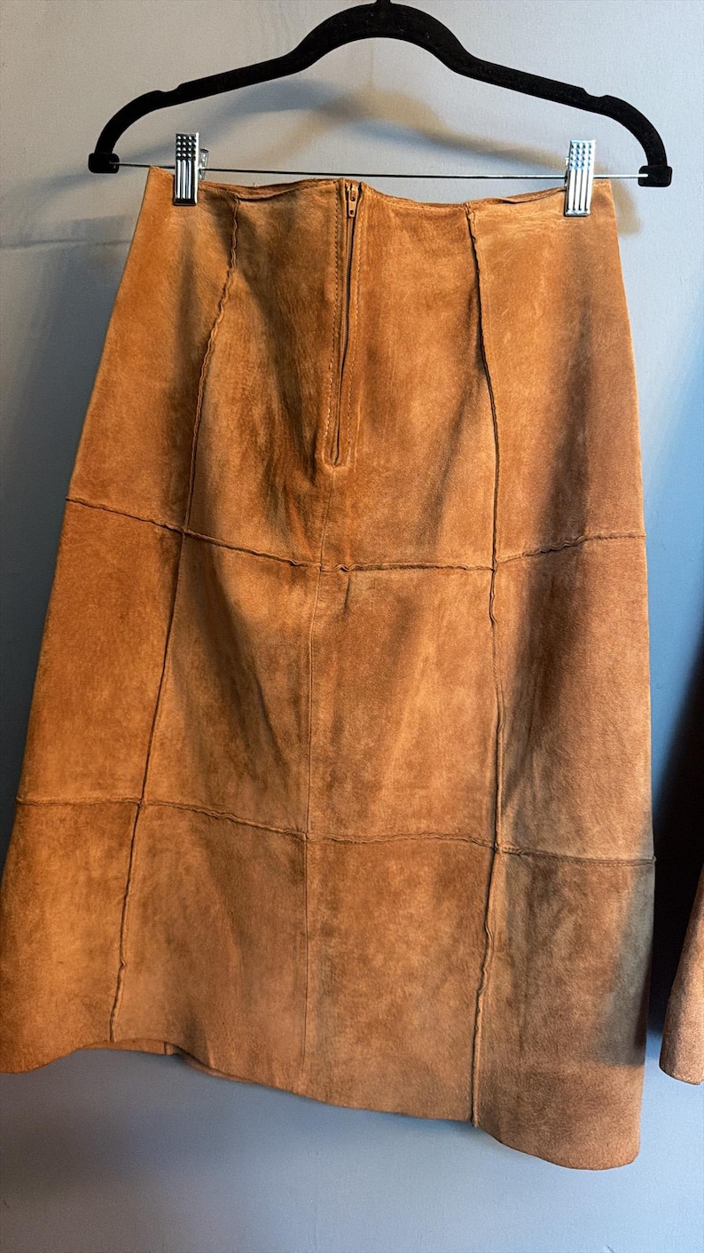 May include: A tan suede skirt featuring a panelled construction with visible vertical and horizontal seams. The skirt has a centred vertical zip closure at the top and is suspended from a black hanger with two metal clips at the waistband.