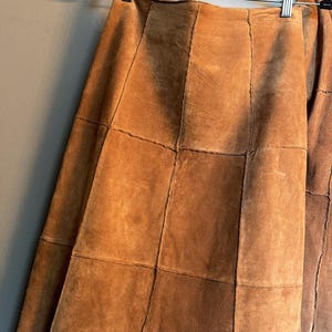 May include: A tan suede skirt constructed from multiple rectangular panels stitched together. The skirt features a high waistline and a straight silhouette, hanging from a black plastic hanger with silver metal clips against a plain wall.
