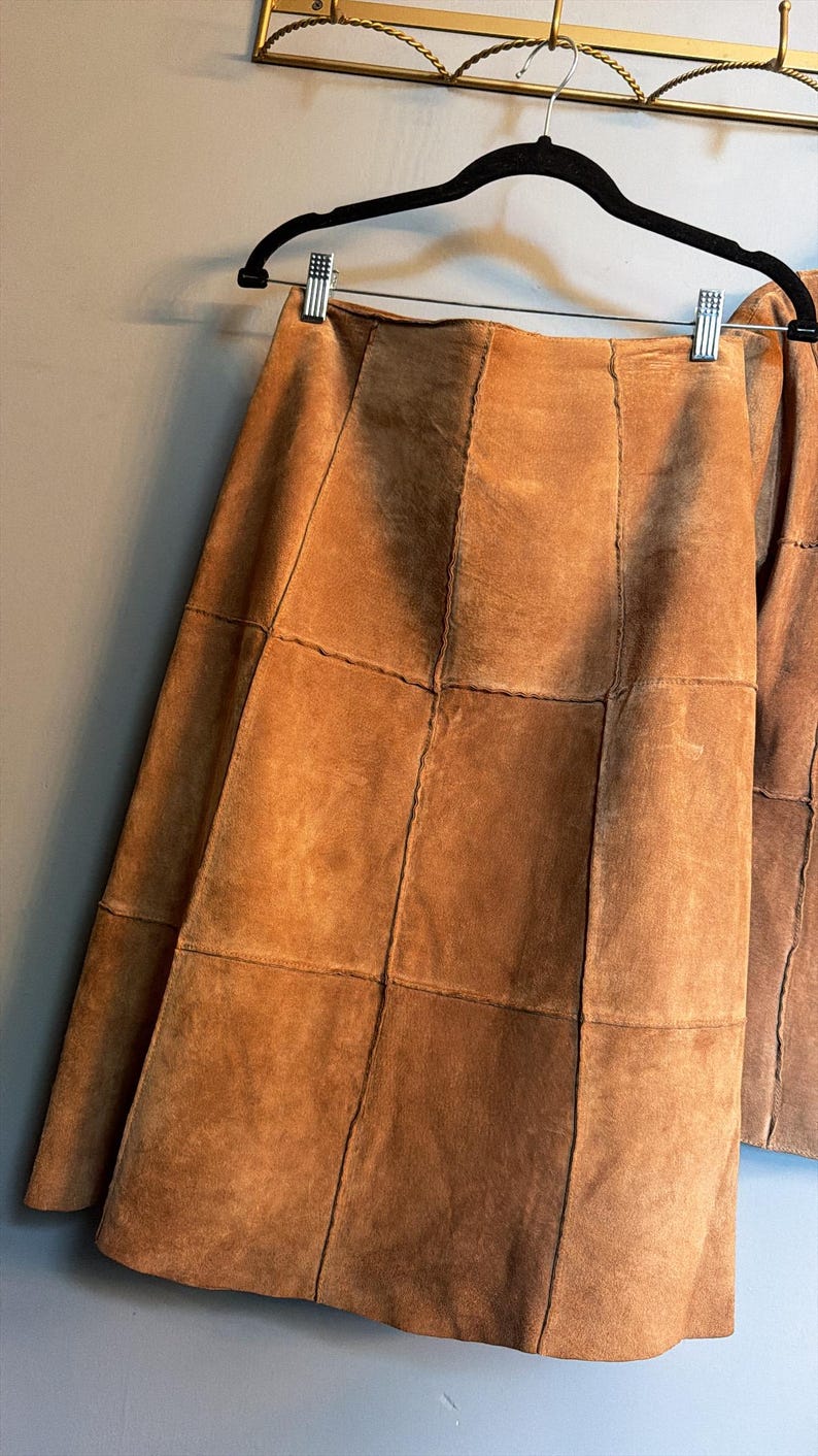 May include: A tan suede skirt constructed from multiple rectangular panels stitched together. The skirt features a high waistline and a straight silhouette, hanging from a black plastic hanger with silver metal clips against a plain wall.