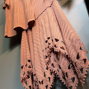 May include: A two-piece peach-colored knit outfit, including a top and a skirt. The top has short sleeves with ruffled edges and decorative elements. The skirt features a unique openwork design with a scalloped hemline. The outfit is hanging on a hanger.
