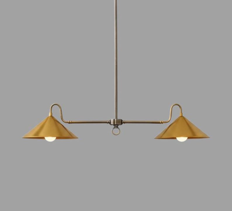 May include: A modern, two-light pendant lamp with a brass finish. The fixture features two conical shades, each with a light bulb, suspended from a central rod and horizontal arm. The design is minimalist and elegant.