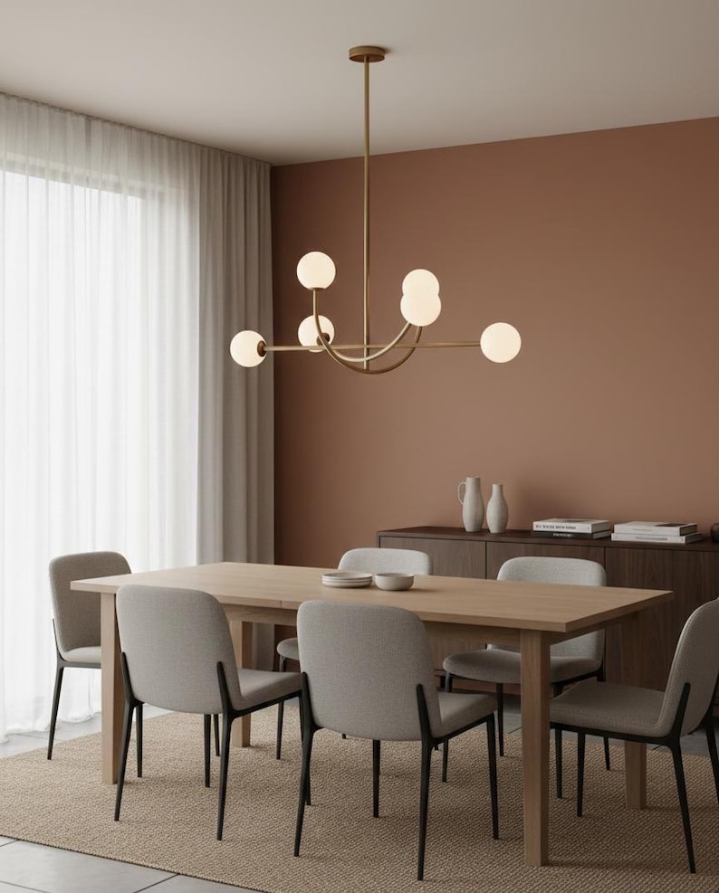 Pu&ograve; includere: Una sala da pranzo moderna con un tavolo in legno chiaro e sei sedie grigie imbottite. Sopra il tavolo &egrave; appeso un lampadario dorato con luci sferiche. La stanza ha una palette di colori neutri con una parete marrone e un tappeto beige.