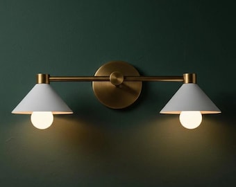 Puede incluir: Una lámpara de pared moderna con dos pantallas cónicas blancas y brazos y placa de montaje de color latón. Cada pantalla tiene una sola bombilla redonda. La lámpara está montada en una pared verde oscuro.