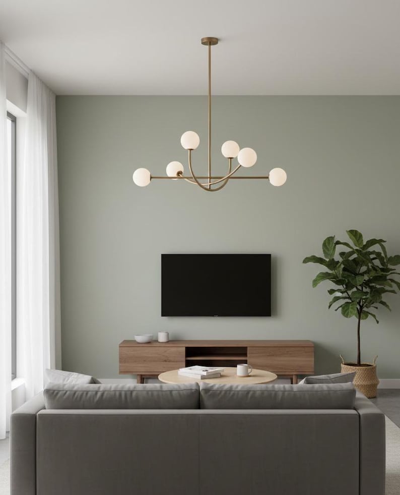 Pu&ograve; includere: Un moderno soggiorno con un divano grigio, un mobile TV in legno e un tavolino rotondo. Un grande televisore nero &egrave; montato a parete. Un lampadario in ottone con luci a globo bianche pende dal soffitto. Una pianta in vaso aggiunge verde.