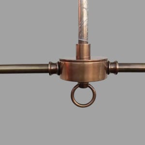 May include: Close-up of a bronze-colored ceiling light fixture. The fixture has a central circular housing with a ring at the bottom and three horizontal arms extending outwards. A vertical rod extends upwards from the center.