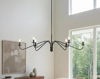 May include: Black metal chandelier with six candle-style lights. The fixture is suspended from the ceiling and hangs above a wooden dining table. The room has a neutral color palette with a large window.