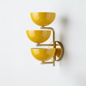 May include: A wall light with three yellow, bowl-shaped shades. The shades are arranged vertically and connected by a brass-coloured metal frame. The fixture is mounted on a round brass-coloured base.