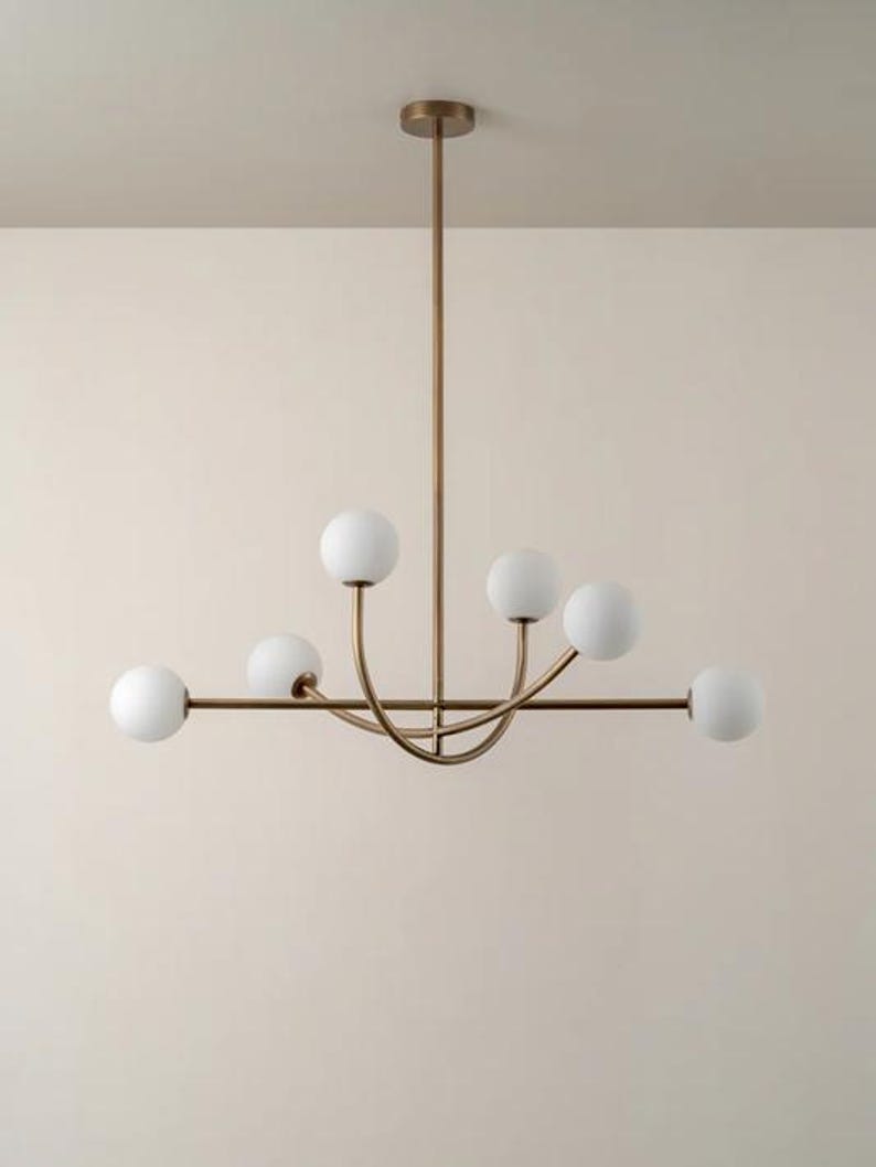 Pu&ograve; includere: Un lampadario moderno con finitura in ottone e sei paralumi sferici in vetro bianco. L'apparecchio &egrave; sospeso al soffitto da un'unica asta. Il design &egrave; minimalista ed elegante, adatto a vari stili di interni.