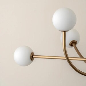 Pu&ograve; includere: Un lampadario moderno con una struttura dorata e bracci curvi. Ogni braccio sostiene un globo di vetro bianco e sferico. L'apparecchio &egrave; progettato con un'estetica minimalista, adatta a vari stili di interni.