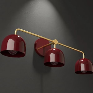 Può includere: Un moderno apparecchio di illuminazione a parete con tre paralumi a cupola color bordeaux. L'apparecchio ha un braccio curvo color oro e un supporto a parete circolare. Le lampadine sono visibili all'interno dei paralumi.