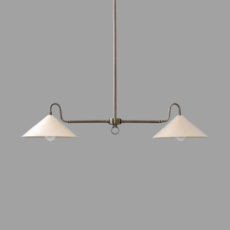 May include: A two-light pendant light fixture with a bronze finish. The fixture has two off-white cone-shaped shades and a central ring detail. The light bulbs are visible within the shades. The fixture is suspended from a single rod.