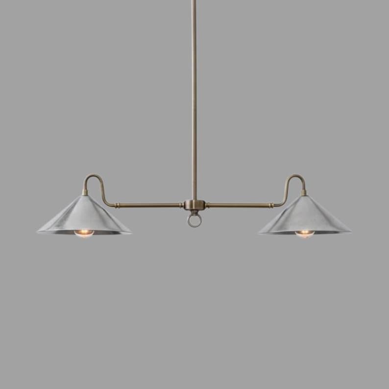 May include: A two-light pendant light fixture with a brushed metal finish. The fixture has a central rod with a horizontal bar extending to two cone-shaped shades. The shades are silver, and the fixture has a warm brass finish.