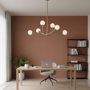 Pu&ograve; includere: Un ufficio con una scrivania in legno chiaro, una sedia girevole grigia e un laptop. Un lampadario dorato con luci sferiche &egrave; appeso sopra. Le pareti sono dipinte di un caldo colore terracotta e una libreria nera &egrave; visibile.
