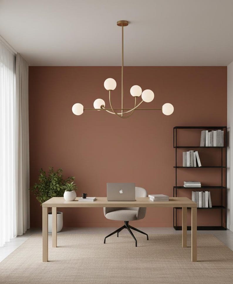 Pu&ograve; includere: Un ufficio con una scrivania in legno chiaro, una sedia girevole grigia e un laptop. Un lampadario dorato con luci sferiche &egrave; appeso sopra. Le pareti sono dipinte di un caldo colore terracotta e una libreria nera &egrave; visibile.