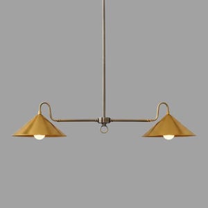 May include: A modern, two-light pendant lamp with a brass finish. The fixture features two conical shades, each with a light bulb, suspended from a central rod and horizontal arm. The design is minimalist and elegant.