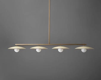 May include: A modern, linear chandelier with four lights. Each light features a white globe bulb and a circular, cream-colored shade. The fixture has a brass finish and hangs from a single vertical rod.