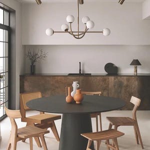 Pu&ograve; includere: Una sala da pranzo moderna con un tavolo rotondo scuro e sedie in legno. Un lampadario in ottone con luci a globo bianche &egrave; appeso sopra il tavolo. Vasi decorativi nei toni del bianco e dell'arancione adornano il tavolo, esaltando l'estetica della stanza.