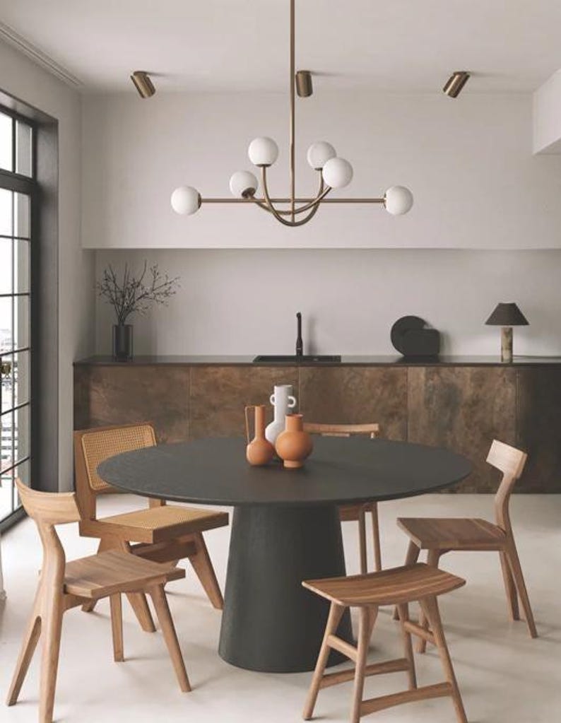 Pu&ograve; includere: Una sala da pranzo moderna con un tavolo rotondo scuro e sedie in legno. Un lampadario in ottone con luci a globo bianche &egrave; appeso sopra il tavolo. Vasi decorativi nei toni del bianco e dell'arancione adornano il tavolo, esaltando l'estetica della stanza.