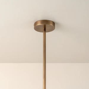 Pu&ograve; includere: Primo piano di un apparecchio di illuminazione a soffitto color bronzo. L'apparecchio ha una base circolare attaccata a un'asta lunga e sottile. L'asta si estende verso il basso dalla base, suggerendo un design minimalista. Sfondo bianco.