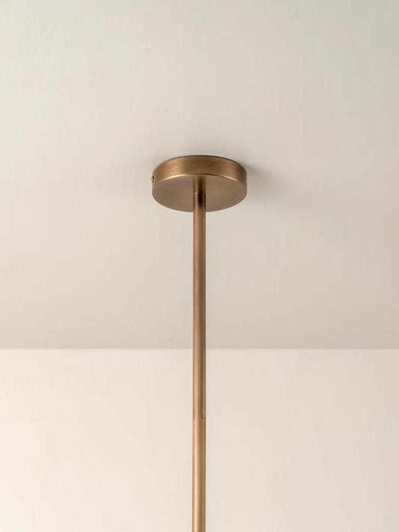 Pu&ograve; includere: Primo piano di un apparecchio di illuminazione a soffitto color bronzo. L'apparecchio ha una base circolare attaccata a un'asta lunga e sottile. L'asta si estende verso il basso dalla base, suggerendo un design minimalista. Sfondo bianco.