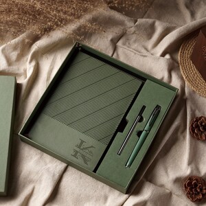 May include: An olive green gift set featuring a notebook and pen. The notebook has diagonal lines and a monogram. The pen is a matching olive green color with a silver-colored pen. The set is presented in a matching olive green box.