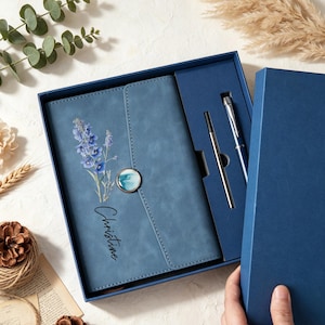 Könnte beinhalten: Ein blaues Notizbuch mit einem Blumenaufdruck aus violetten Ritterspornblüten und dem Namen Christine in schwarzer Schreibschrift. Das Notizbuch hat einen runden, marmorierten Knopfverschluss und wird in einer blauen Geschenkbox mit passendem Stift geliefert.