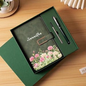 May include: A dark green notebook with the name Samantha printed in white, featuring a floral print of pink and cream carnations at the bottom and a gold-toned metal clasp. The set includes a matching dark green pen and a silver ink refill.