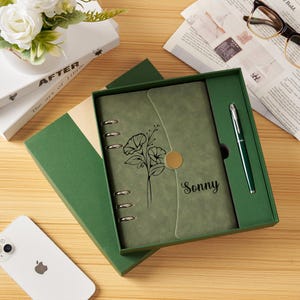 Può includere: Un diario in similpelle verde salvia con meccanismo a sei anelli, caratterizzato da un'illustrazione floreale a linee nere e dal nome Sonny stampato sulla copertina. Il diario include una penna verde abbinata ed è presentato in una scatola.