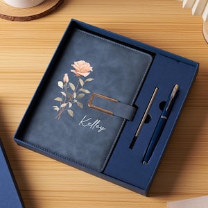 Peut inclure: Un ensemble de journal et de stylo bleus dans une boîte cadeau. Le journal présente un motif floral et le nom "Kelley". L'ensemble comprend un stylo bleu assorti et un stylo argenté. Le journal a une fermeture à boucle.