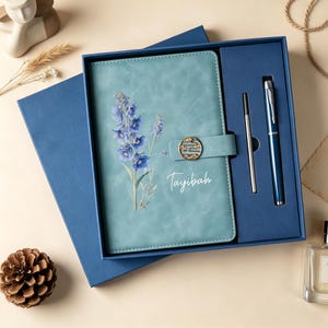 Puede incluir: Una caja de regalo azul que contiene un cuaderno azul claro con estampado floral, el nombre Tayibah en letra blanca y un cierre circular dorado. Un bolígrafo azul a juego y un recambio de tinta plateado están en un compartimento lateral.