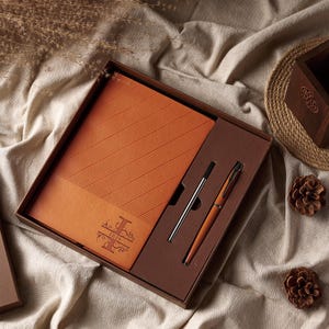 May include: A gift set featuring a leather-bound notebook and two pens, presented in a brown box. The notebook is orange with diagonal lines and the word "IRIS" in a decorative font. One pen is silver, the other is orange.