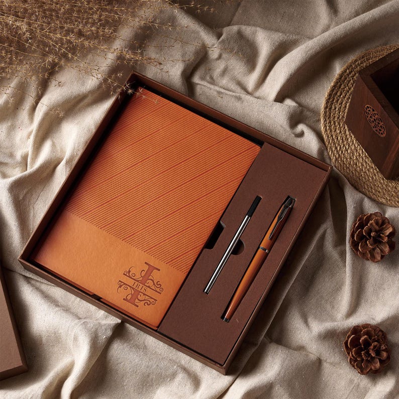 May include: A gift set featuring a leather-bound notebook and two pens, presented in a brown box. The notebook is orange with diagonal lines and the word "IRIS" in a decorative font. One pen is silver, the other is orange.