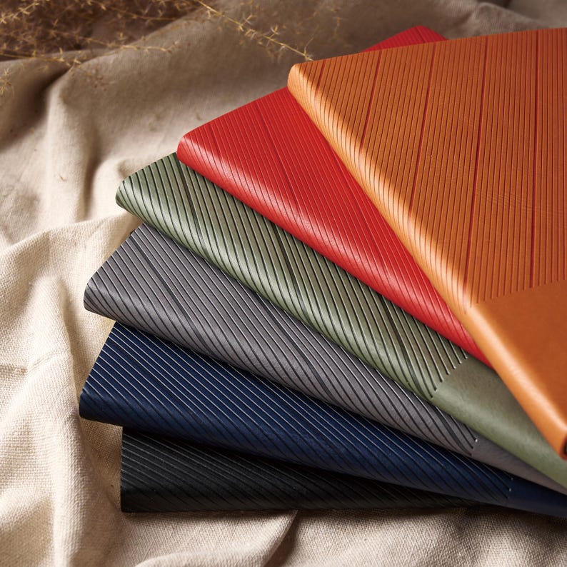 May include: A stack of colorful, textured notebooks. The notebooks are in shades of red, orange, green, gray, navy, and black. Each notebook has a diagonal striped pattern on the cover.