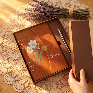 Puede incluir: Un juego de diario de cuero marrón con un bolígrafo, presentado en una caja de regalo. El diario presenta un diseño floral y el nombre "Dina" en escritura cursiva. Un ramo de lavanda se encuentra encima de la caja sobre un tapete de encaje.