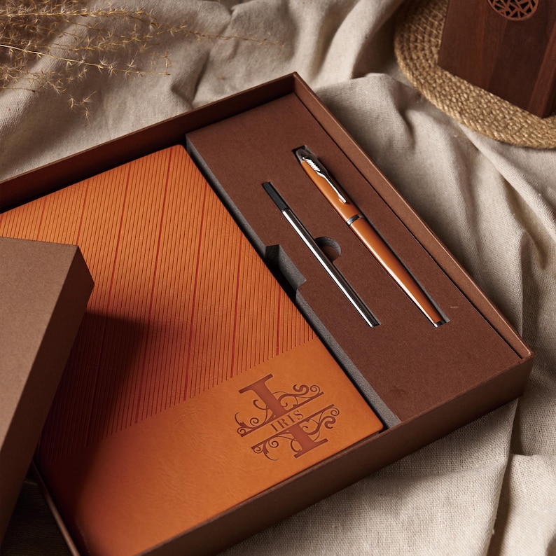 May include: A boxed gift set featuring a leather-bound notebook and a pen. The notebook is orange with vertical lines and the name "IRIS" is embossed on the cover. The pen is orange and silver. The box is brown.