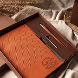 May include: A boxed gift set featuring a burnt orange leather-bound journal with vertical lines and the word "Queen" embossed on the lower right corner. A matching pen and refill are nestled in the box. The pen has a silver tip and a burnt orange barrel.